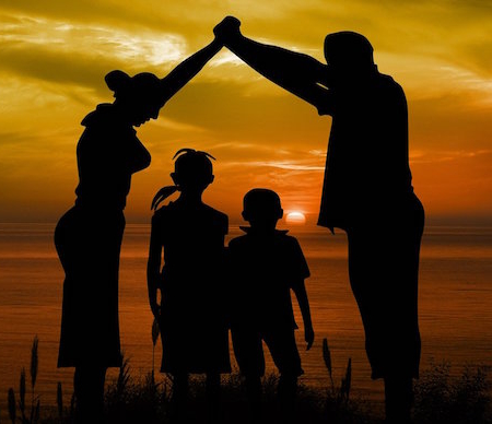 Shows an image of a family on the beach at sunset. A mother and father are holding their hands together creating an umbrella of protection above the heads of their children.