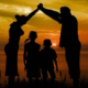 Shows an image of a family on the beach at sunset. A mother and father are holding their hands together creating an umbrella of protection above the heads of their children.