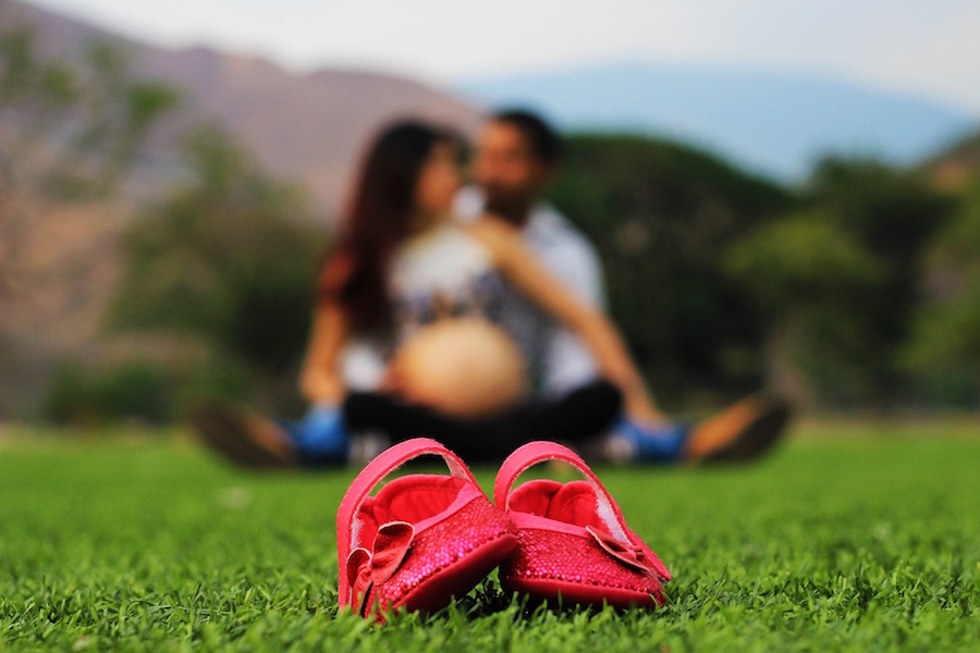 Shows a pair of red baby shoes withan expectant couple sitting on the grass in the background