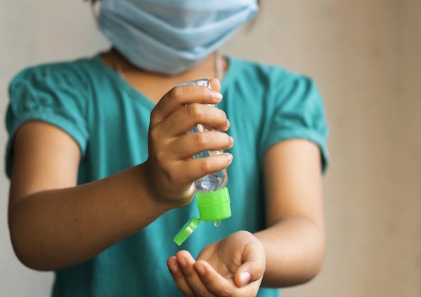 Shows an image of a small female child wearing a mask and squeezing hand sanitizer into her hand