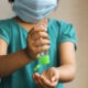 Shows an image of a small female child wearing a mask and squeezing hand sanitizer into her hand