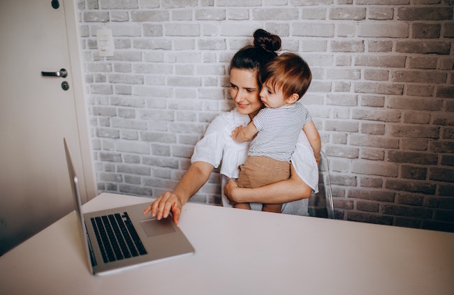 Shows a woman at a desk holding an infant while she is trying to type on a laptop