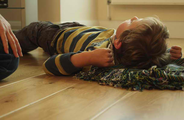 Shows a young boy lying on the floor in the aftermath of a seizure. A woman's hand resting on her leg can be seen to the left of him.