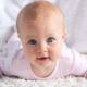 Shows a baby girl with bright blue eyes in a pink onesie laying on her stomach on top of a white shag rug. She is holding her head up and smiling while looking directly at the camera.