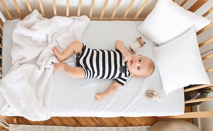 Image shows a baby on its back in a crib surrounded by blanket, pillows, and toys