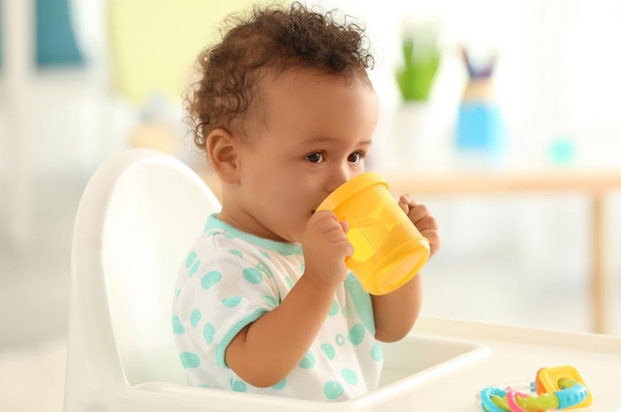 Shows a baby boy in a high chair drinking from a yellow sippy cup