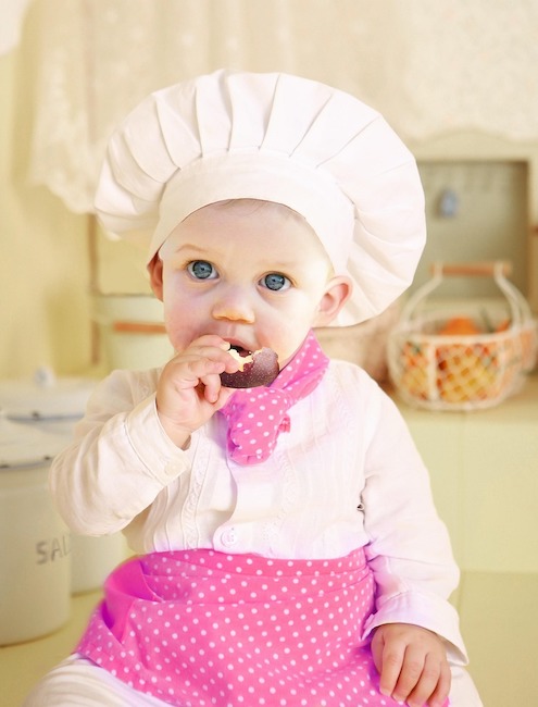 Shows a baby girl eating a piece of apple while sittiing on a counter wearing chef whites with a pink scarf and a pink skirt.