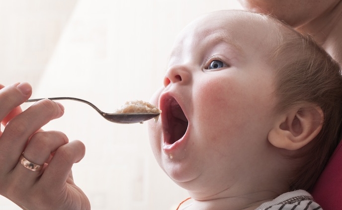 Shows a baby with its mouth wide open being spoon fed