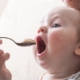 Shows a baby with its mouth wide open being spoon fed