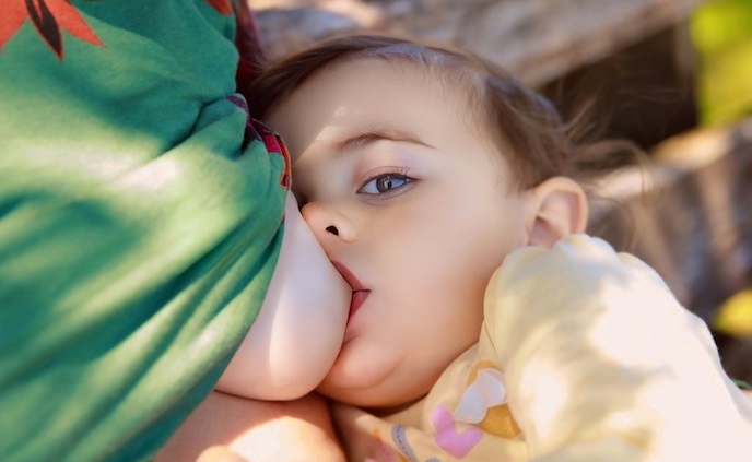 The image shows a baby in a yellow outfit breastfeeding from her mother's breast