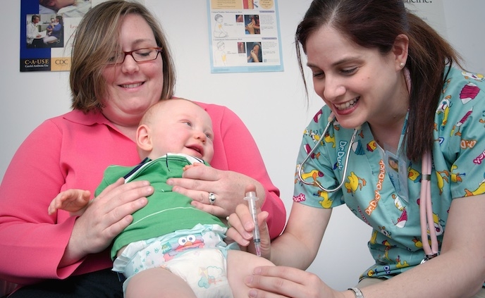 Shows a nurse administering a shot to a baby who is being held in its mother's arms.