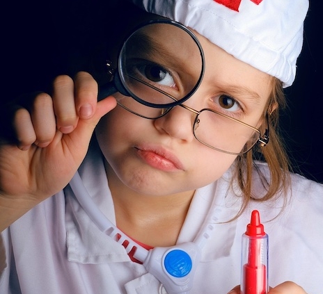 Shows a young girl wearing glasses dressed as a nurse. She is holding a magnifying glass in one hand and a plastic play syringe in the other.