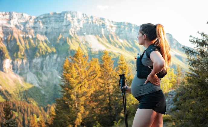 Shows a pregnant woman holding a walking stick as she stands on the peak of a mountain